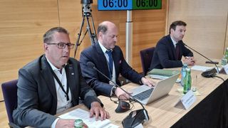Three men in suits sit at a conference table, with one using a laptop, another looking at papers, and the third using a microphone. The room has wooden walls and a digital clock above.