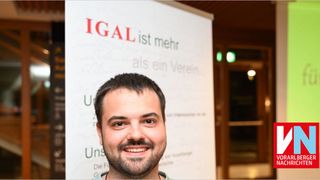 A man with a beard smiles in front of a banner that says IGAL is more than a club.