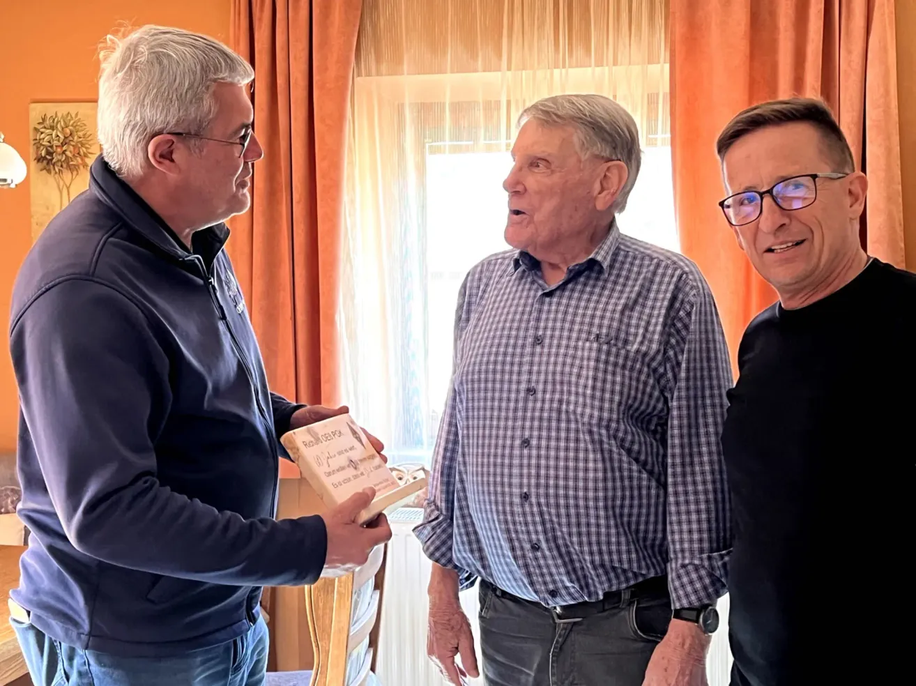 Three men are standing in a room. The man on the left is holding a box and looking at the man in the middle. The man in the middle is smiling and looking at the man on the right. The man on the right is smiling and looking at the camera.