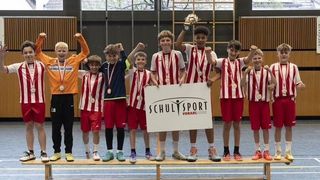 Young athletes in sports uniforms pose with medals and a trophy on a wooden bench in an indoor sports facility.