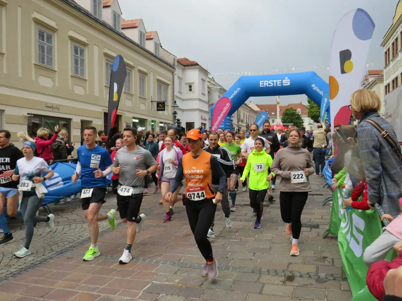 Participants in a marathon are running on a cobblestone street. One runner wears a bright orange vest with the number 9744. In the background, a blue arch with the text 'erste' is visible.
