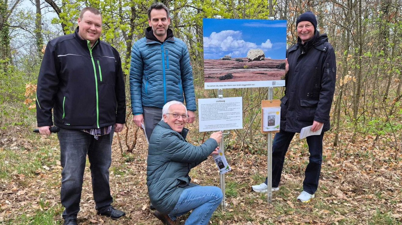 Fünf Männer stehen in einem bewaldeten Gebiet in der Nähe eines Schildes. Ein Mann kniet, um das Schild zu lesen. Ein anderer Mann hält ein Dokument.