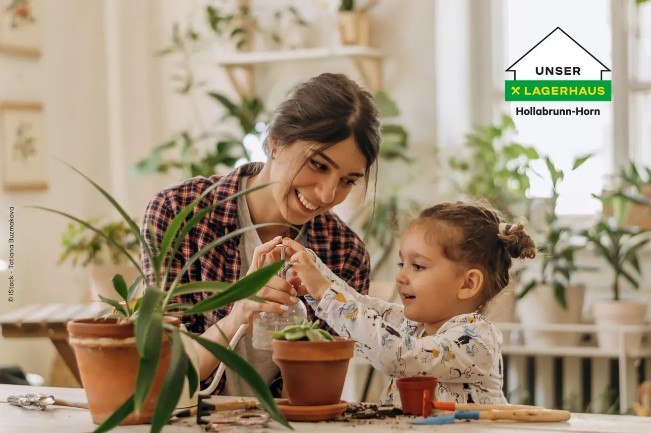 Eine Frau und ein kleines Mädchen pflanzen eine Pflanze in einen Topf, umgeben von anderen Topfpflanzen. Die Frau lächelt und das Mädchen sieht konzentriert aus.