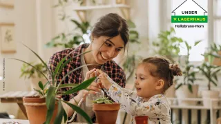 Eine Frau und ein kleines Mädchen pflanzen eine Pflanze in einen Topf, umgeben von anderen Topfpflanzen. Die Frau lächelt und das Mädchen sieht konzentriert aus.