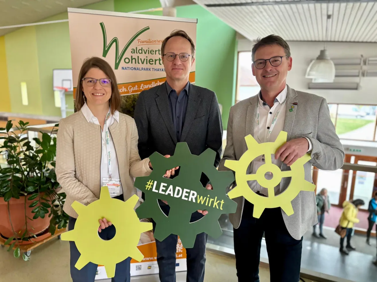 Drei Personen in formeller Kleidung halten gelbe Zahnräder mit '#LEADERwirkt' darauf hoch und lächeln in einem grünen Raum. Hinter ihnen steht ein Banner für 'Alviert ohlviert Nationalpark'.