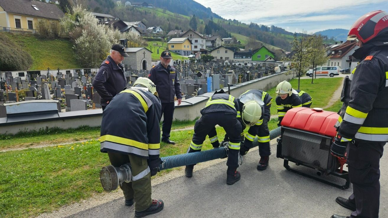 Feuerwehrleute arbeiten mit Schläuchen auf einem Friedhof. Zwei Feuerwehrleute sind gebeugt, zwei andere stehen in der Nähe und ein fünfter beobachtet.