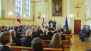 Eine Veranstaltung in einer Kirche. Zwei Personen stehen an einem Rednerpult, möglicherweise halten sie eine Rede. Das Publikum sitzt auf Kirchenbänken, und Flaggen sind an der Wand angebracht.