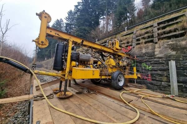 A yellow drilling machine is on a wooden platform near a train track, with trees and a stone wall in the background.