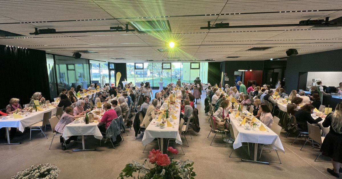 Eine große Indoor-Essveranstaltung mit Tischen, die mit weißen Tüchern, verschiedenen Blumen und Tellern mit Essen bedeckt sind. Gäste sitzen um die Tische herum, manche tragen Masken.