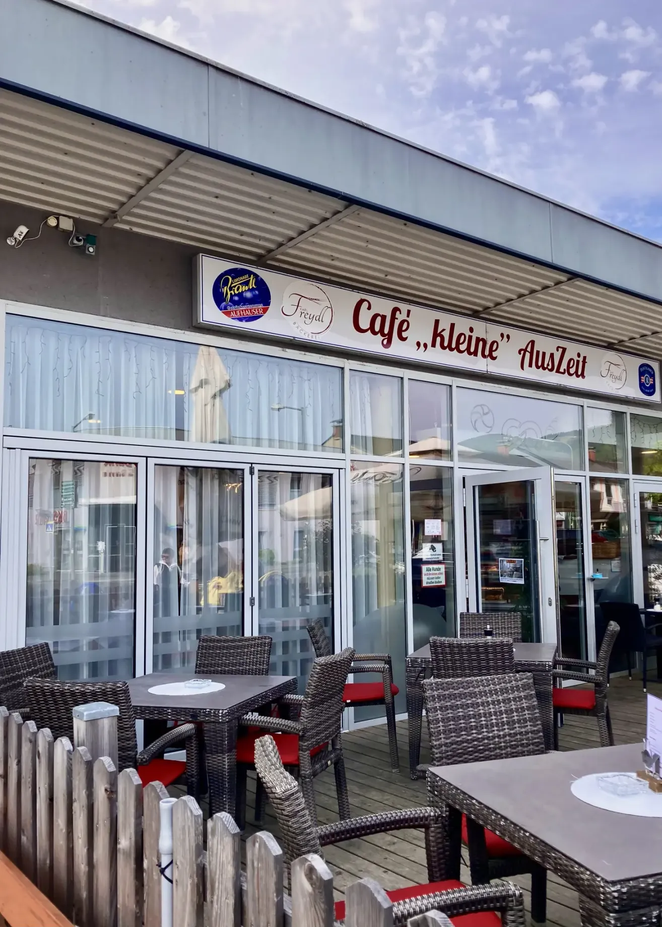 Die Vorderseite eines Cafes mit Glastüren und Fenstern. Das Schild trägt die Aufschrift 'Café,,kleine' Auszeit'. Außerhalb sind Tische und Stühler aufgestellt.