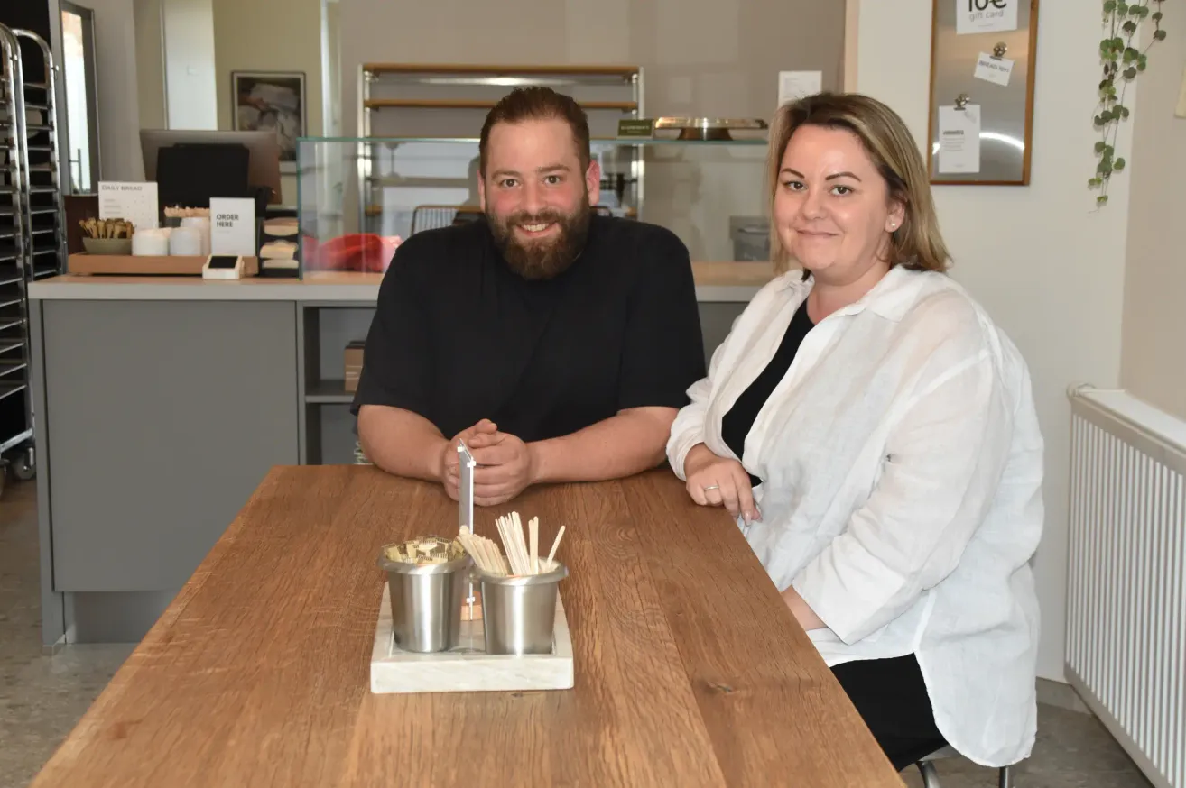 Ein Mann und eine Frau sitzen lächelnd an einem Holztisch. Sie sind von verschiedenen Gegenständen umgeben, darunter zwei Becher mit Chopsticks. Dahinter steht eine Glasvitrine.