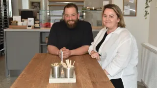 Ein Mann und eine Frau sitzen lächelnd an einem Holztisch. Sie sind von verschiedenen Gegenständen umgeben, darunter zwei Becher mit Chopsticks. Dahinter steht eine Glasvitrine.