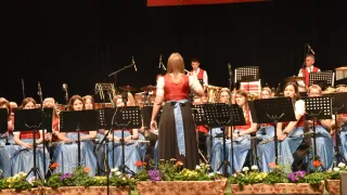 Eine Frau steht in der Mitte auf der Bühne mit einer Band hinter ihr, alle in traditioneller Kleidung gekleidet. Ein Banner mit der Aufschrift 'Musikverein Jugendkapelle Großpetersdorf' hängt über der Bühne.