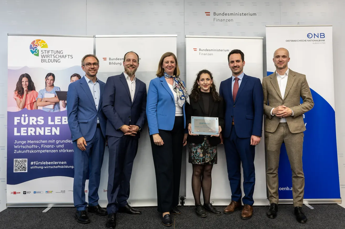 Fünf formell gekleidete Personen stehen vor Bannern bei einer Veranstaltung. Eine Frau in der Mitte hält eine Auszeichnung. Zwei Männer und zwei Frauen stehen neben ihr.