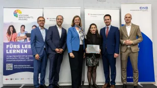 Fünf formell gekleidete Personen stehen vor Bannern bei einer Veranstaltung. Eine Frau in der Mitte hält eine Auszeichnung. Zwei Männer und zwei Frauen stehen neben ihr.