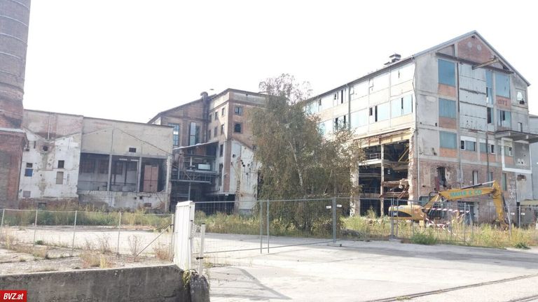 Ein altes Industriegebäude im Bauzustand ist eingezäunt. Ein gelbes Baufahrzeug ist vor dem Zaun geparkt.