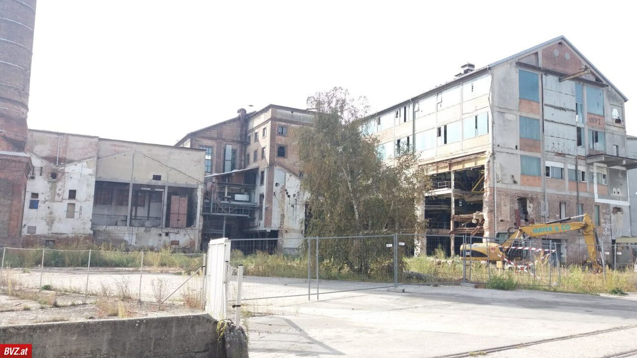 Ein altes Industriegebäude im Bauzustand ist eingezäunt. Ein gelbes Baufahrzeug ist vor dem Zaun geparkt.