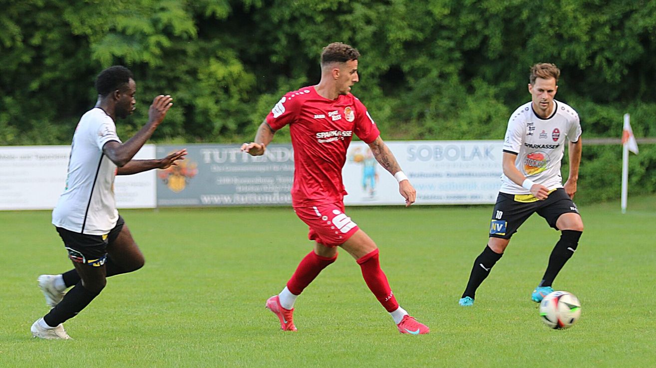 Ein Fußballspieler in einem roten Trikot läuft auf dem Feld. Dahinter ist ein anderer Spieler in Weiß und Schwarz zu sehen. Der Hintergrund zeigt ein grünes Feld mit Bäumen.