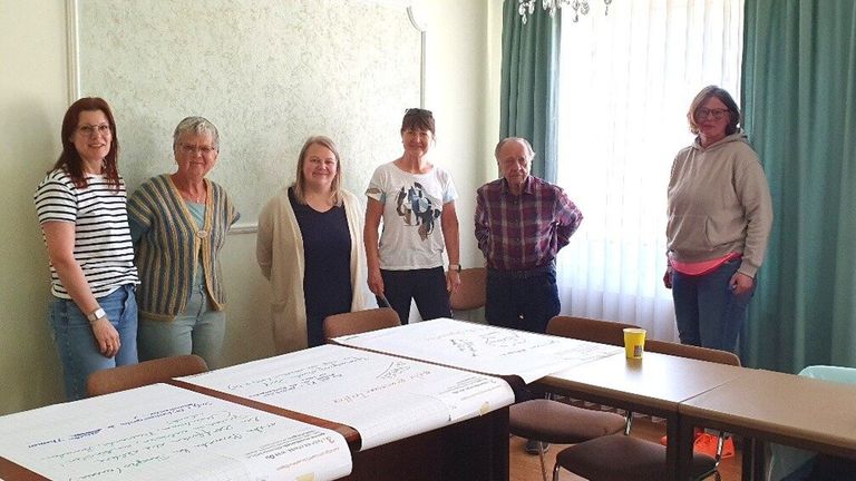 Eine Gruppe von fünf Personen, möglicherweise in einer Besprechung, steht hinter einem Tisch mit Papieren und einem gelben Becher darauf. Sie befinden sich in einem Raum mit weißen Wänden und Fenstern.