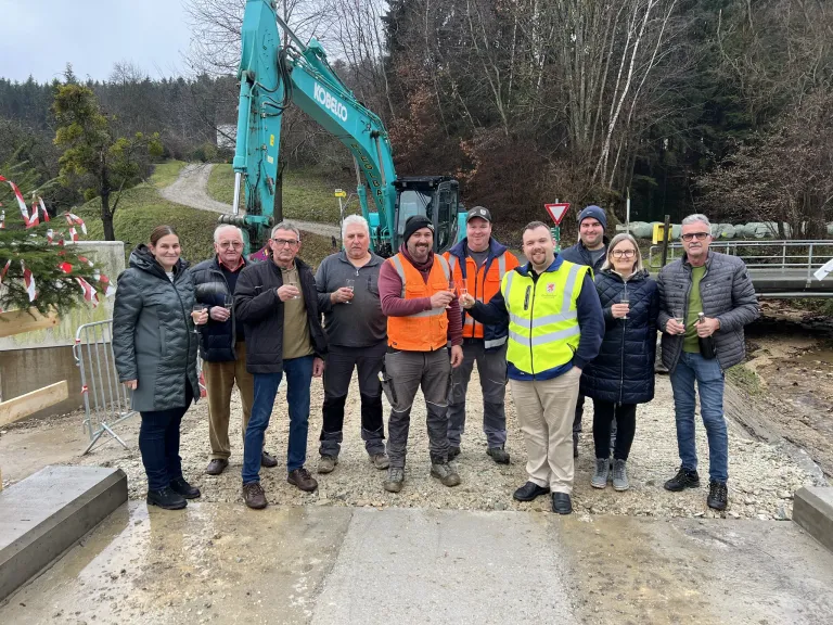 Eine Gruppe von Menschen steht in der Nähe einer Baustelle mit einem grünen Bagger im Hintergrund. Sie halten Weingläser in den Händen und posieren für ein Foto.