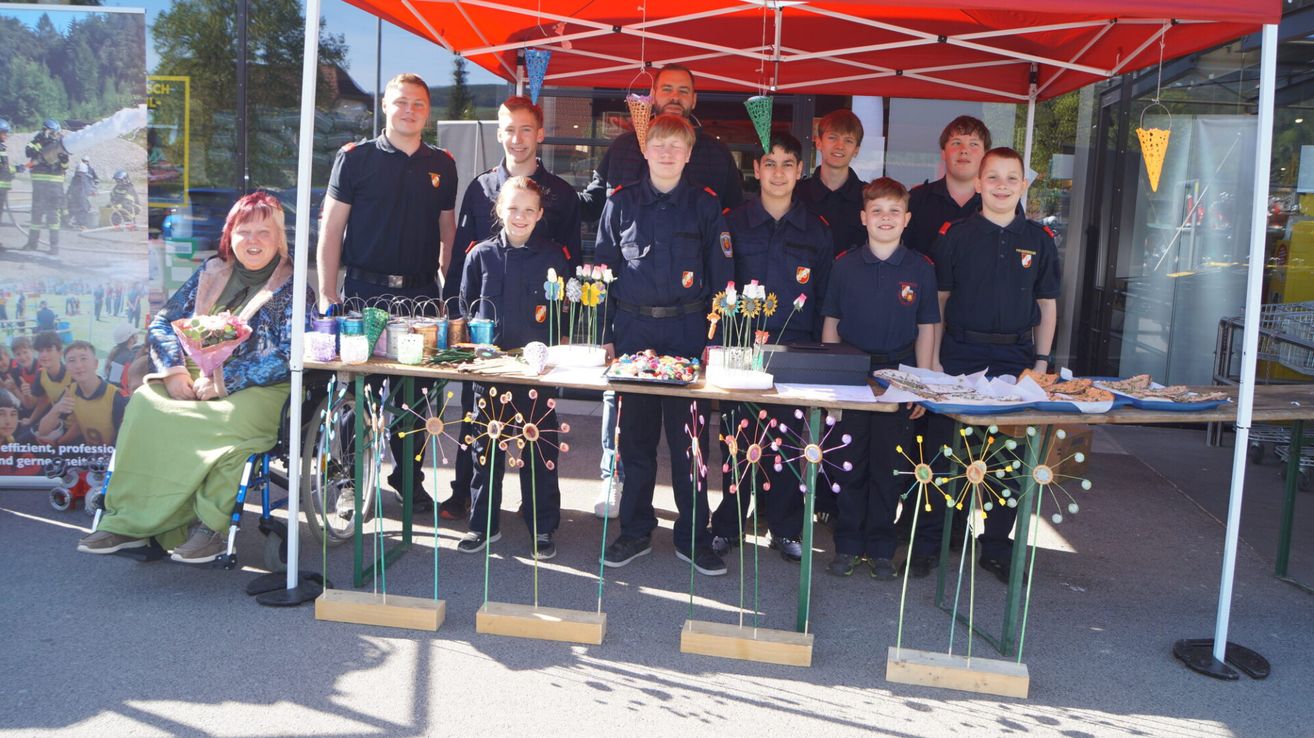 Eine Gruppe junger Feuerwehrleute, darunter auch Mädchen, steht hinter einem Tisch mit bunten Blumen und anderen Gegenständen unter einem roten Zelt. Sie tragen ihre Uniformen.