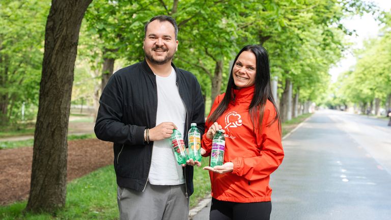 Ein Mann und eine Frau stehen auf einem Weg und halten Flaschen mit Getränken. Sie lächeln und scheinen für ein Foto zu posieren. Hinter ihnen sind Bäume und Gras.