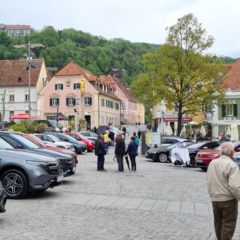 Eine Straßenszene mit Kopfsteinpflaster und geparkten Autos. Mehrere Menschen gehen im Vordergrund. Gebäude säumen die Straße, eines mit einer goldenen Statue. Bäume und ein Hügel sind im Hintergrund.