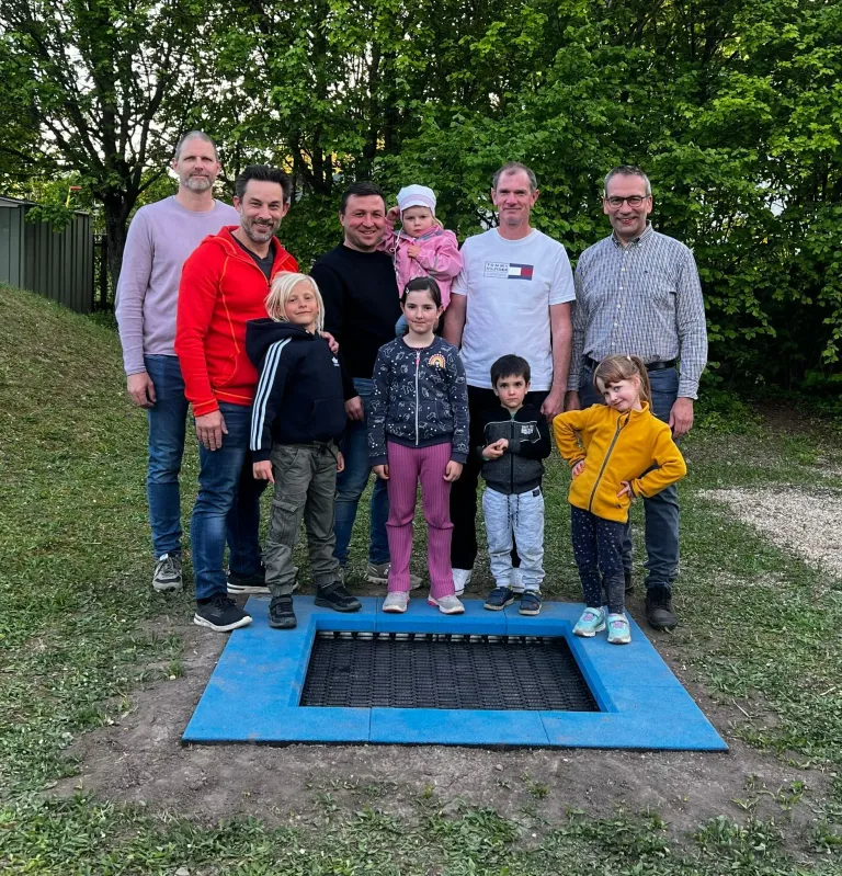 Eine Gruppe von Menschen steht um eine blaue Matte und lächelt für ein Foto. Ein Kind hält ein Baby. Bäume und ein Zaun sind im Hintergrund zu sehen.