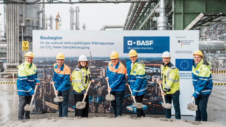 Fünf Personen in Warnwesten und Schutzhelmen halten Schaufeln in der Hand und stehen vor einem großen BASF-Banner auf einer Baustelle.