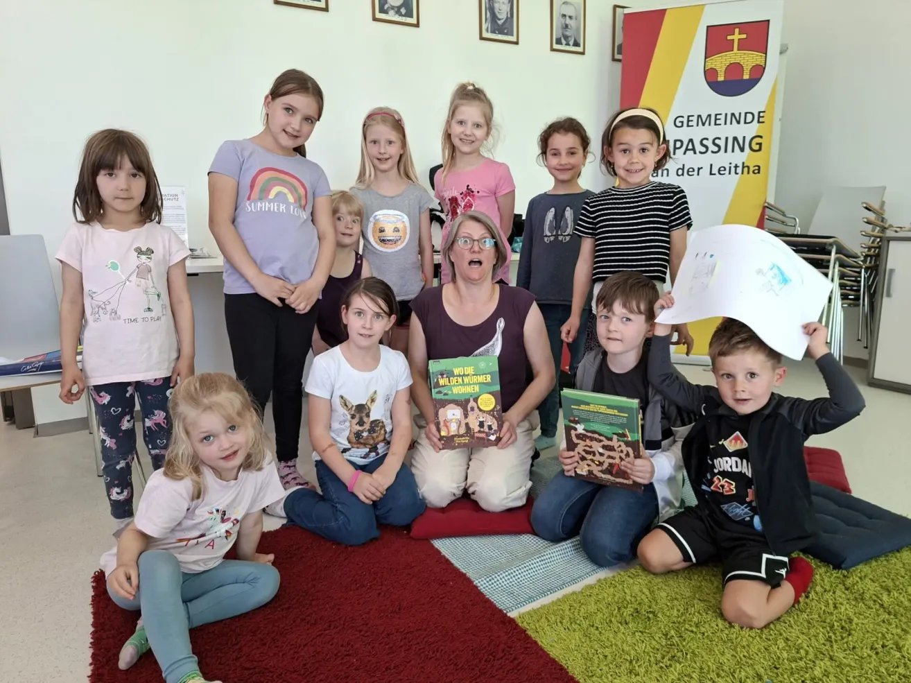 Eine Gruppe von Kindern und einer erwachsenen Frau sitzen mit Büchern im Klassenzimmer auf dem Boden. Die Frau hält ein Buch mit dem Titel 'Wilde Die Wilden Wurmer Von'. Die Kinder lächeln und posieren für ein Foto. Dahinter ist eine Wand mit Bilderrahmen.