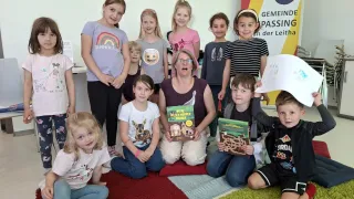 Eine Gruppe von Kindern und einer erwachsenen Frau sitzen mit Büchern im Klassenzimmer auf dem Boden. Die Frau hält ein Buch mit dem Titel 'Wilde Die Wilden Wurmer Von'. Die Kinder lächeln und posieren für ein Foto. Dahinter ist eine Wand mit Bilderrahmen.