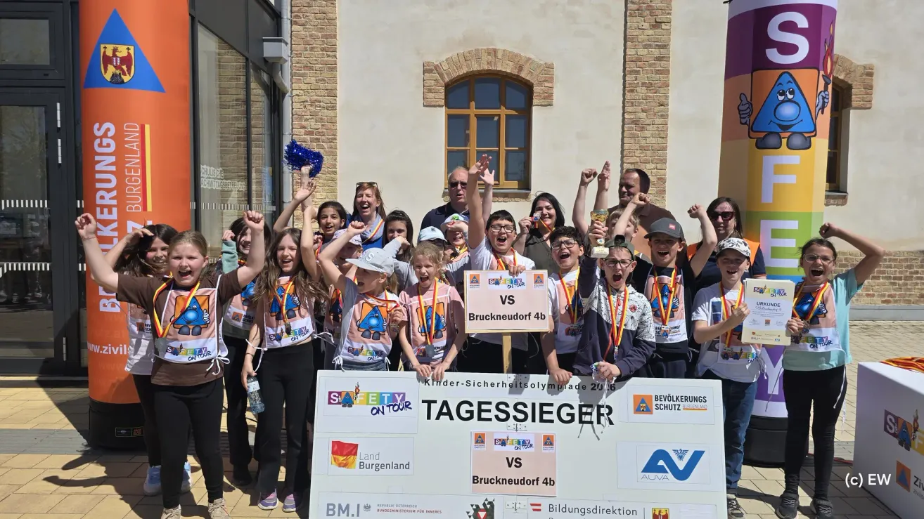 Eine Gruppe von Kindern und Erwachsenen steht zusammen, hält ein großes Schild und jubelt. Sie tragen passende Uniformen und einige haben Medaillen. Das Schild sagt Bruckneudorf 4b und Sicherheit auf Tour.