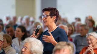 Eine ältere Frau spricht in ein Mikrofon in einem überfüllten Raum. Sie trägt eine Brille und eine Halskette. Das Publikum ist im Hintergrund verschwommen.