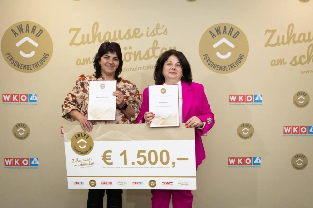 Zwei Frauen stehen hinter einem Schild mit €1.500 und halten Urkunden in den Händen. Der Hintergrund zeigt ein Logo und Text. Sie lächeln.
