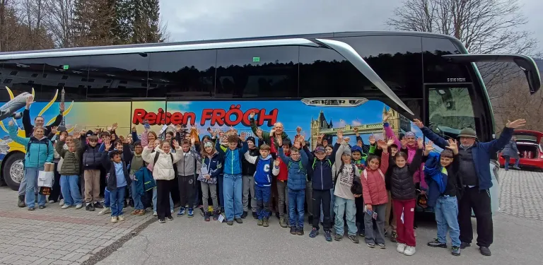 Eine Gruppe von Kindern steht vor einem Bus mit einem farbenfrohen Design und 'Reisen FRÖCH' darauf geschrieben. Sie winken mit den Händen und lächeln.