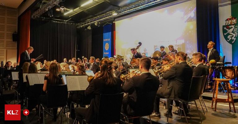Ein großes Orchester spielt auf der Bühne mit einer großen Leinwand im Hintergrund. Die Musiker spielen verschiedene Instrumente, und einige sitzen.