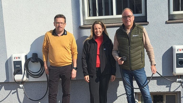 Three people stand outside a house, smiling. A man on the left wears a yellow shirt, the middle woman a black jacket over a red top. The man on the right wears a green vest and holds a black cable.