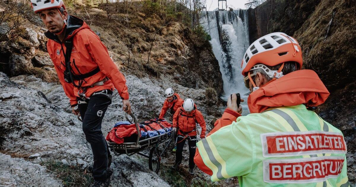 Rettungskräfte tragen eine Trage in der Nähe eines Wasserfalls. Sie sind mit Helmen und orangefarbenen Jacken ausgestattet. Einer hält eine Kamera.