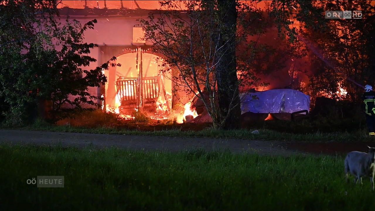 Ein Haus steht in Flammen, wobei sich das Feuer auf die nahe gelegenen Bäume ausbreitet. Das Gebäude ist schwer beschädigt und Rauch erfüllt die Luft.