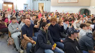 Ein überfüllter Raum mit Personen, wahrscheinlich Studenten, die rote Karten hochhalten und nach oben schauen. Einige sitzen, andere stehen. Eine Person vorne trägt eine Brille und eine blaue Jacke.