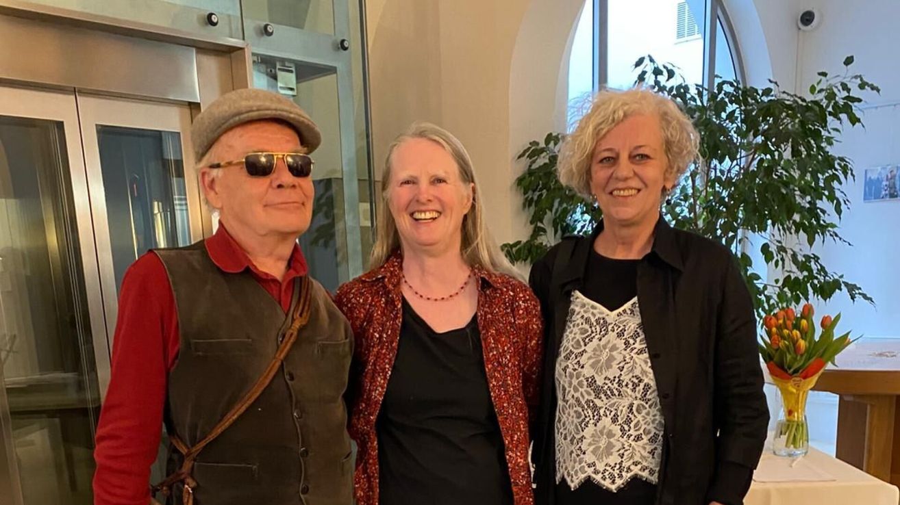 Drei Personen, zwei Frauen und ein Mann, lächeln und posieren für ein Foto vor einem Gebäude. Die Frauen tragen schwarze Oberteile und Jacken.