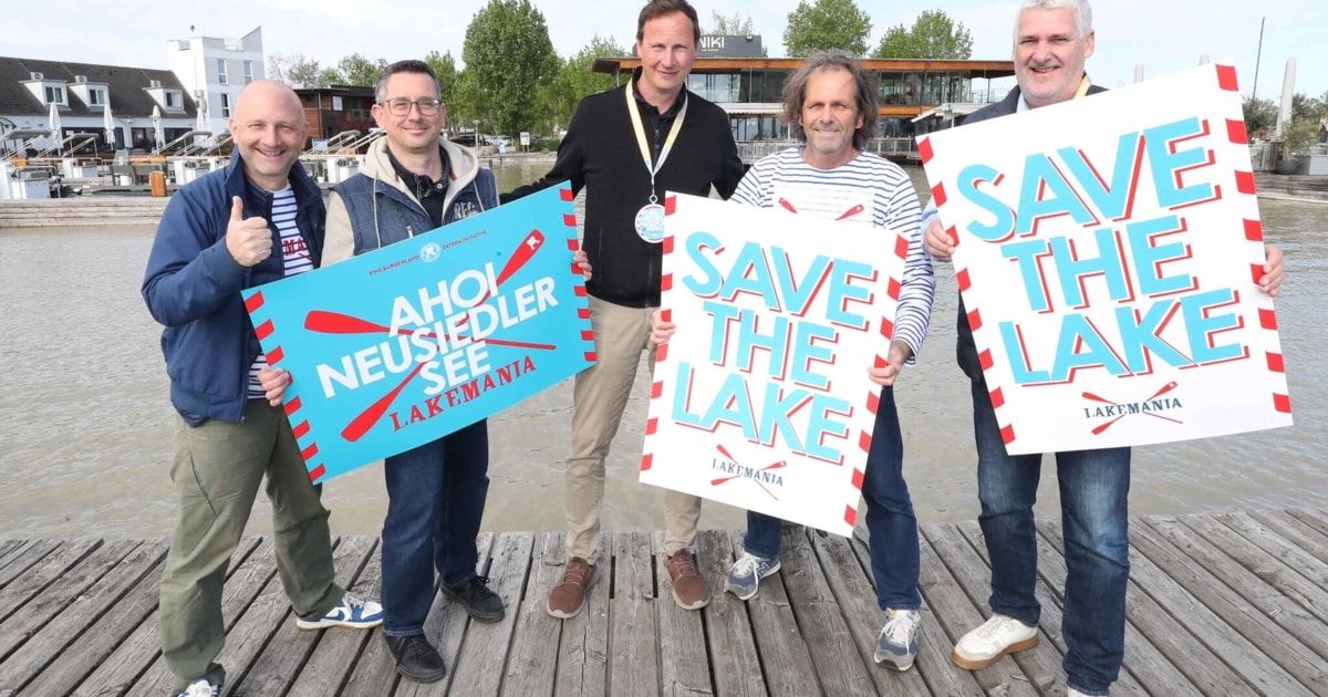 Fünf Männer stehen auf einem Steg und halten Schilder mit 'Save the Lake' und 'Neusiedler See Lakemania'. Einer trägt eine Medaille. Hinter ihnen ein See und Gebäude.