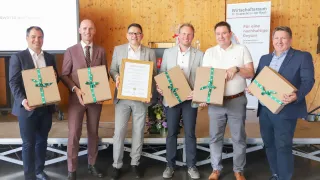 Fünf Männer in Anzügen halten Geschenke und Urkunden und lächeln in einem Raum mit einer Holzwand und Blumen. Ein Schild mit weißem Text ist im Hintergrund zu sehen.
