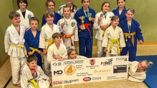 Gruppenfoto von jungen Judokas in blauen und weißen Uniformen mit verschiedenen Medaillen, die mit einer Trophäe für ein Foto posieren.
