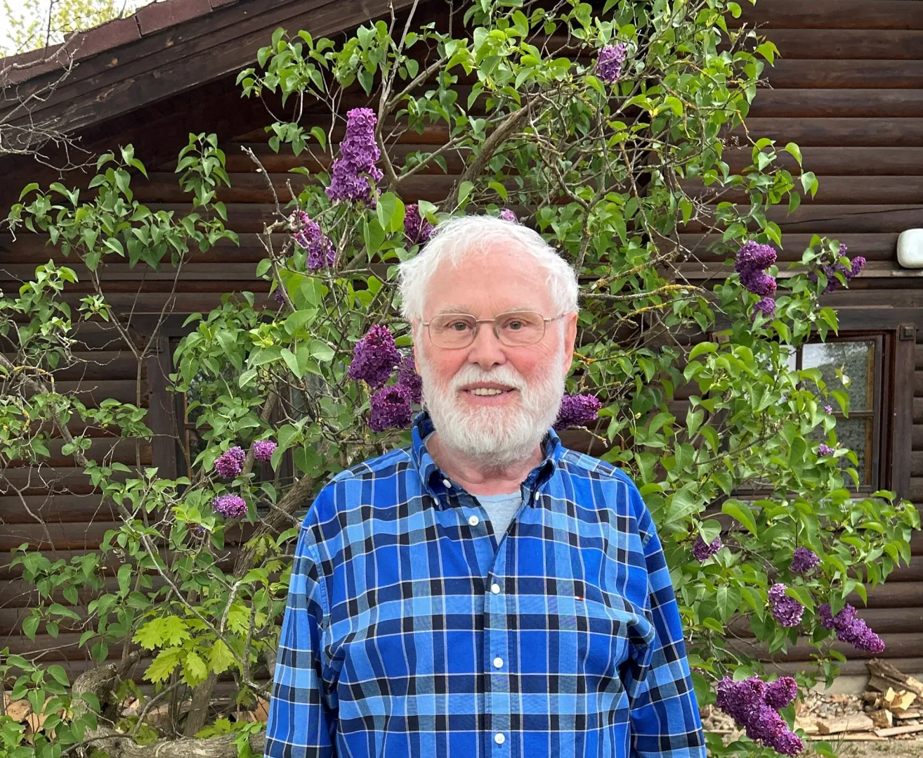 Ein Mann mit weißem Haar und Bart steht vor einem Gebäude mit einem Baum mit lila Blüten. Er trägt ein blaues kariertes Hemd.