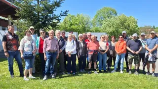 Eine Gruppe von Menschen steht auf einem Rasen, lächelt und posiert für ein Foto. Sie tragen Freizeitkleidung. Dahinter steht ein großer Baum und einige Bäume in der Ferne.