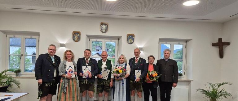 Eine Gruppe von Menschen in traditioneller bayerischer Kleidung, die Auszeichnungen und Blumen halten, steht in einem Raum mit Blick auf die Berge.