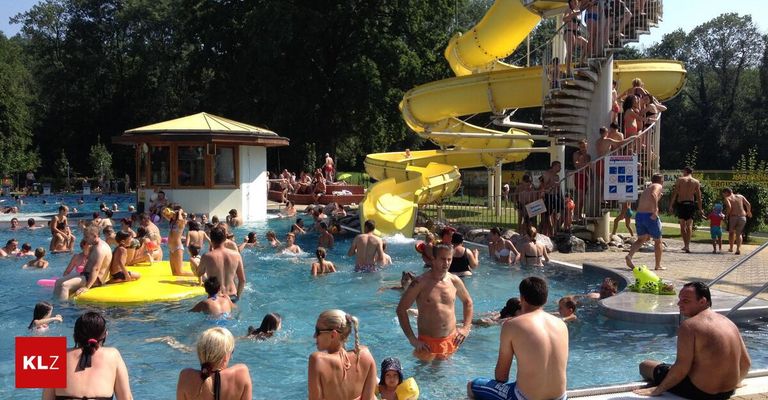 Menschen schwimmen im Pool und genießen die gelbe Wasserrutsche. Ein Gebäude mit gelbem Dach steht in der Nähe des Pools. Einige Leute sitzen auf den Stufen der Wasserrutsche.