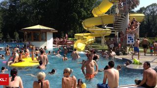 Menschen schwimmen im Pool und genießen die gelbe Wasserrutsche. Ein Gebäude mit gelbem Dach steht in der Nähe des Pools. Einige Leute sitzen auf den Stufen der Wasserrutsche.