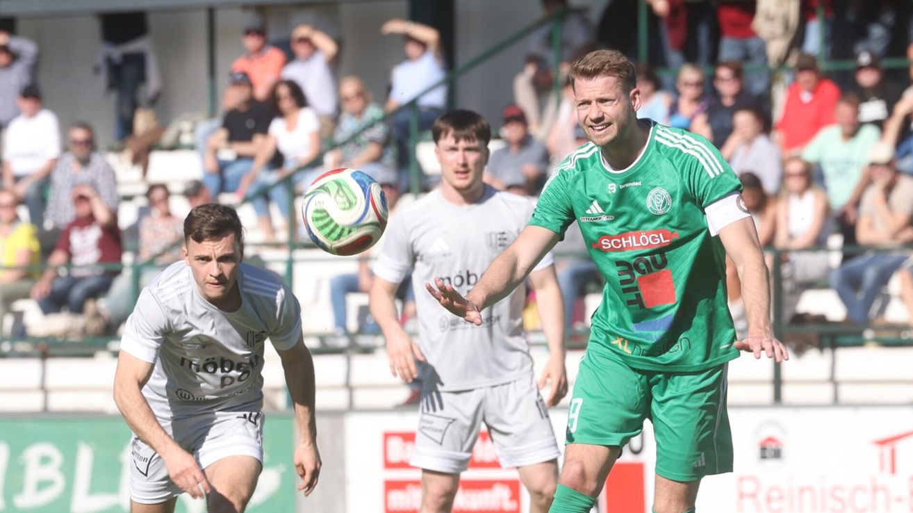 Drei Fußballspieler auf dem Feld. Einer in grünem Trikot zielt darauf ab, den Ball zu treten. Zuschauer beobachten von den Tribünen.